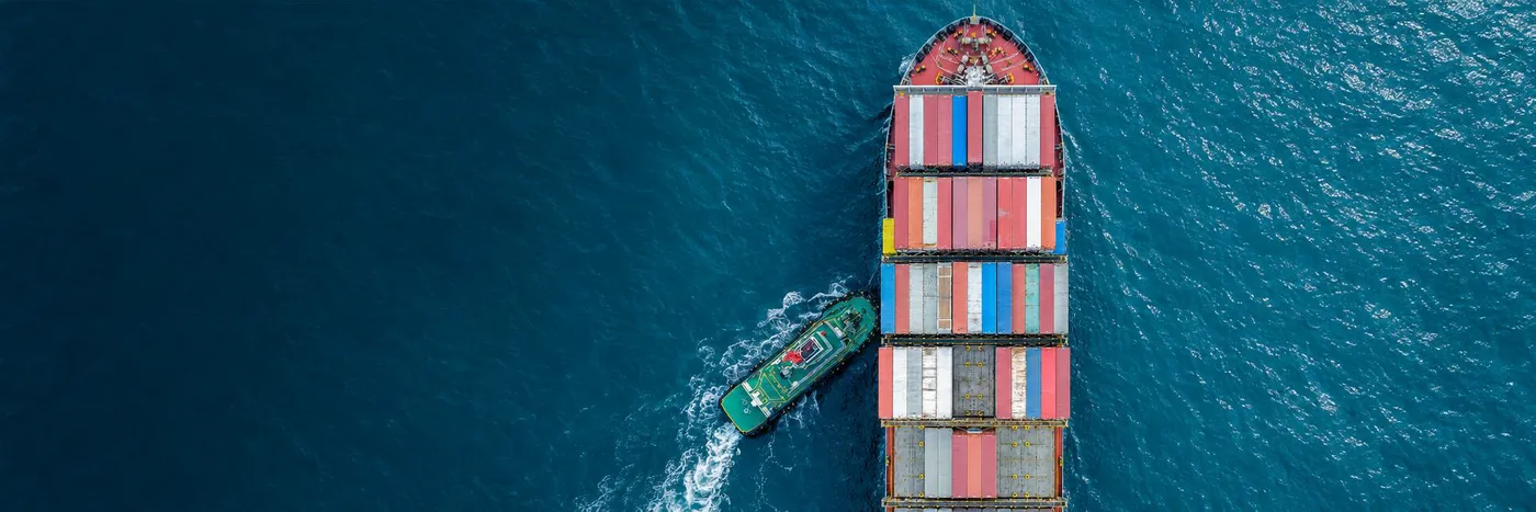 Aerial view of a loaded container ship assisted by a green tugboat on deep teal-blue water, darker open water on the left
